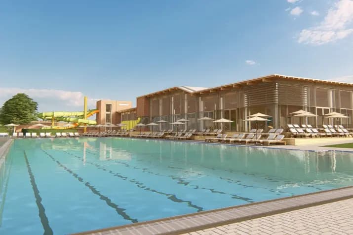 Ein moderner Außenpool, umgeben von Liegestühlen und Sonnenschirmen, mit einer Wasserrutsche im Hintergrund. Gebäude mit großen Fenstern sind neben dem Pool sichtbar.